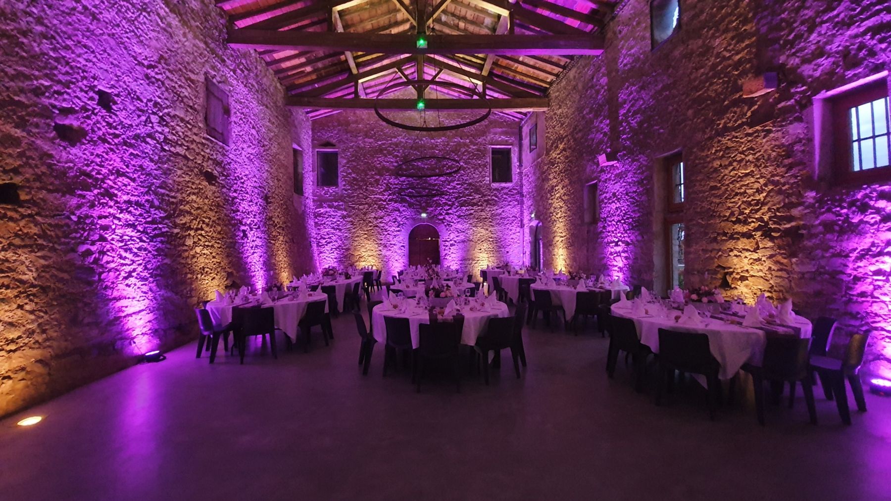 Salle de réception aux murs de pierre baignée de lumière violette, tables dressées pour un mariage